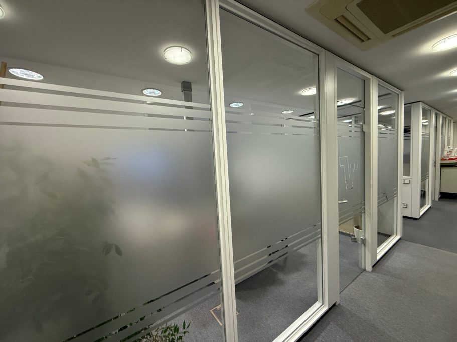 Büroflur mit Glastüren und matten Fenstern, dahinter Pflanzen sichtbar.