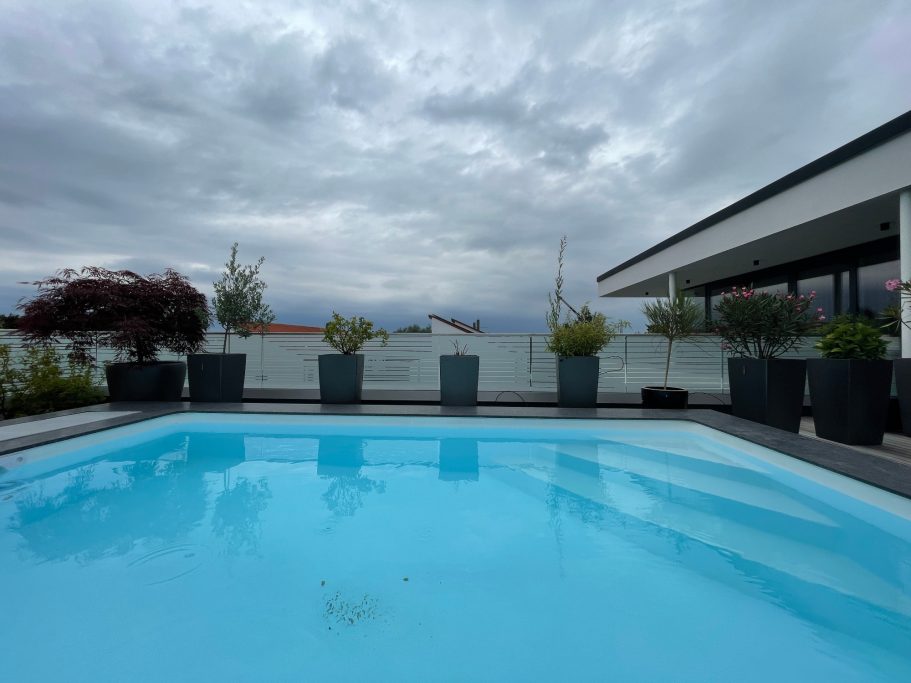 Blauer Pool umgeben von hohen Pflanzkübeln und einem bewölkten Himmel.