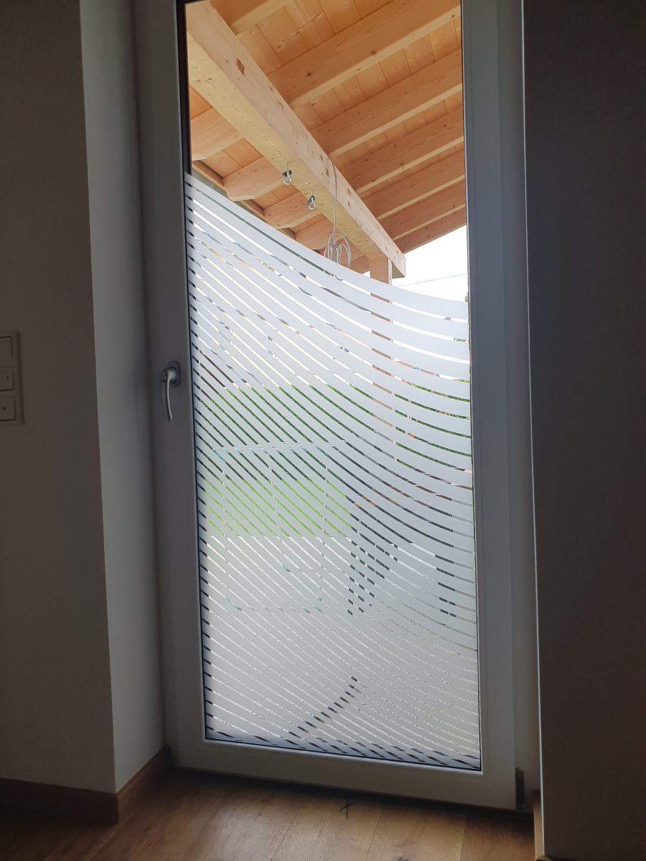 Glastür mit dekorativem Muster, Blick auf eine Terrasse und einen Garten im Hintergrund.