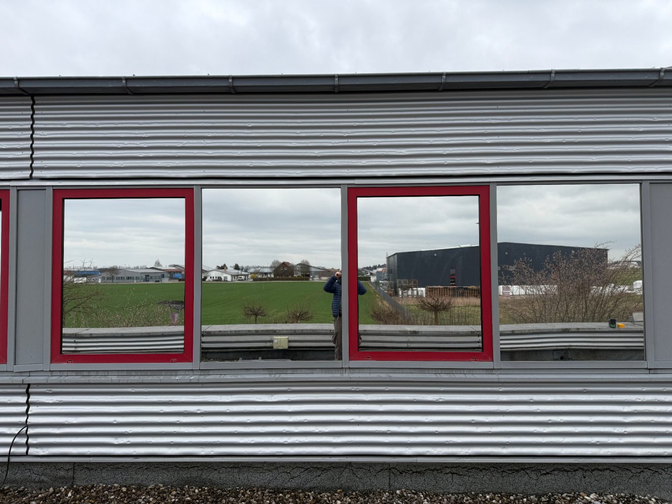 Drei rote Fensterrahmen spiegeln eine graue Landschaft und einen dunkelgrauen Gebäudekomplex wider.