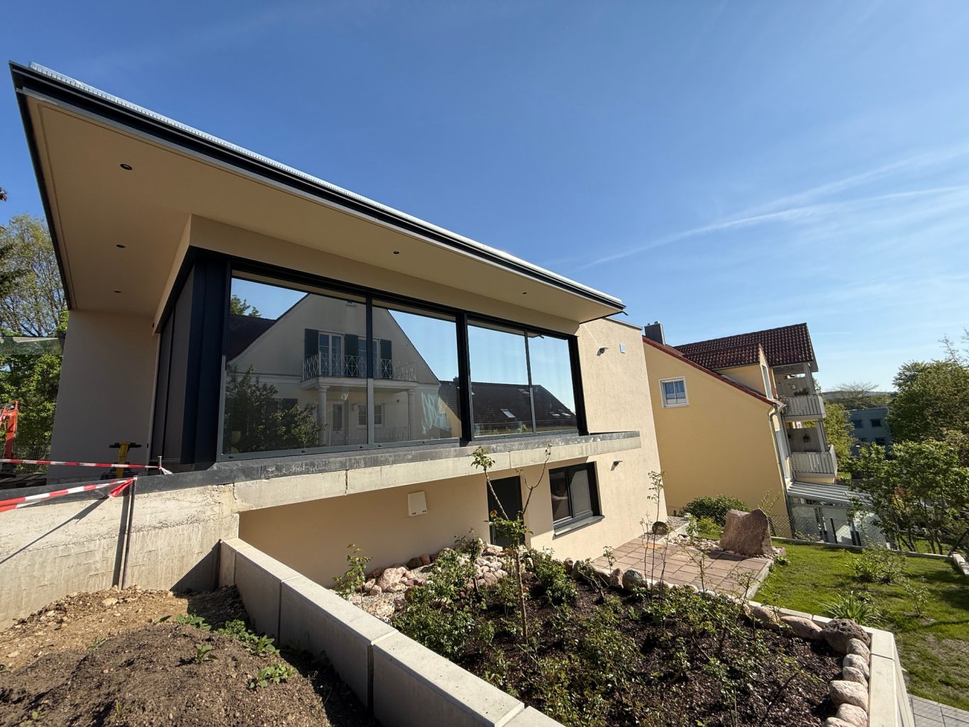 Moderner Hausbau mit großen Fenstern, umgeben von Garten und blauer Himmel.