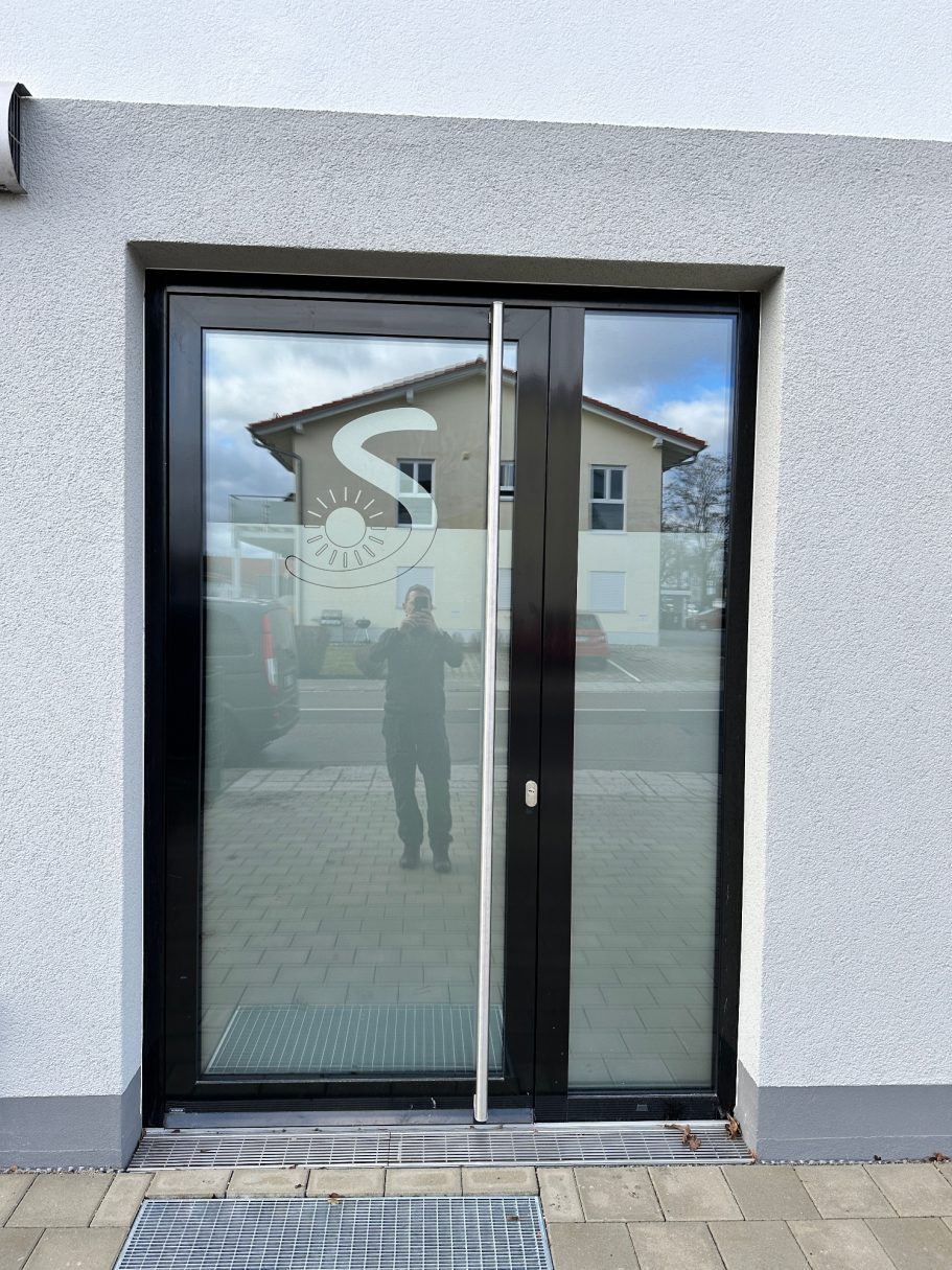 Moderne Glastür mit schwarzem Rahmen, reflektierender Oberfläche und grauem Hintergrund.