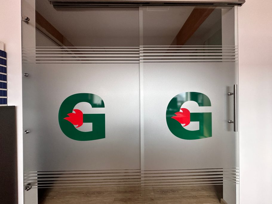 Glas-Schiebetür mit zwei großen, grünen "G"-Buchstaben und rotem Pfeil.