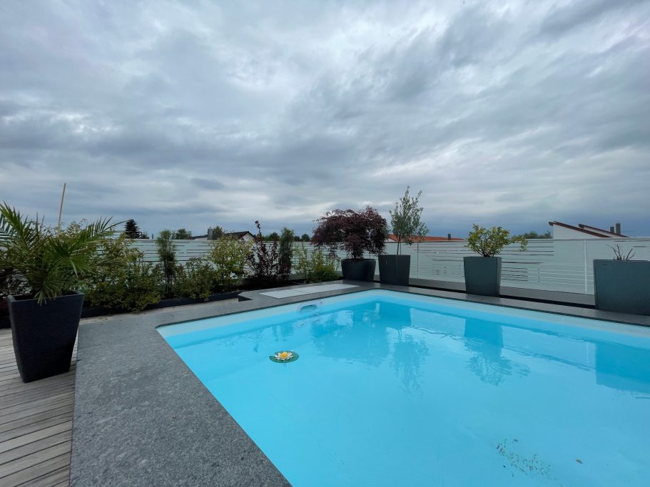 Moderner Schwimmingpool auf einer Dachterrasse, umgeben von Pflanzen unter einem bewölkten Himmel.